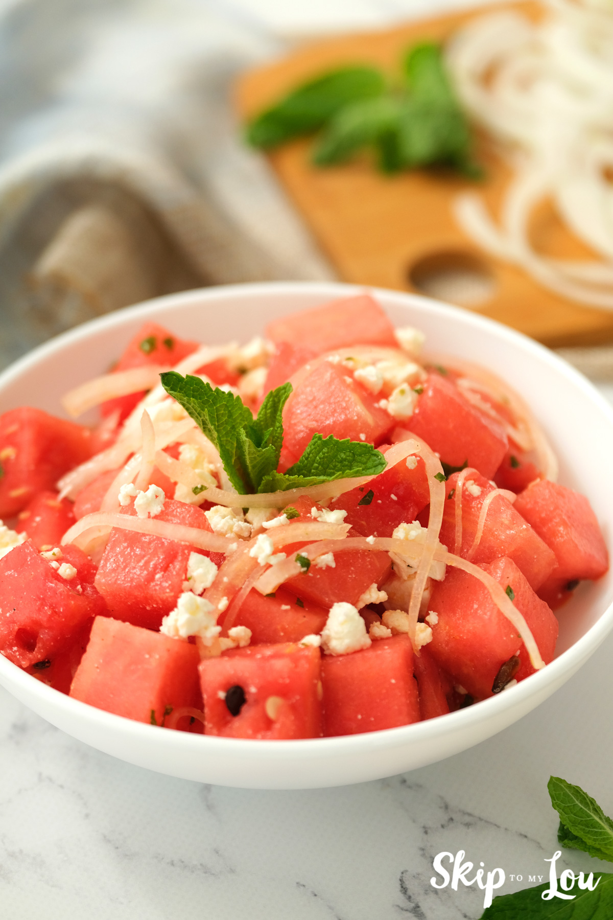 Cubes of red watermelon, red onion slices, crumbled cheese, a sprig of green mint in a white serving bowl, by Skip to my Lou.