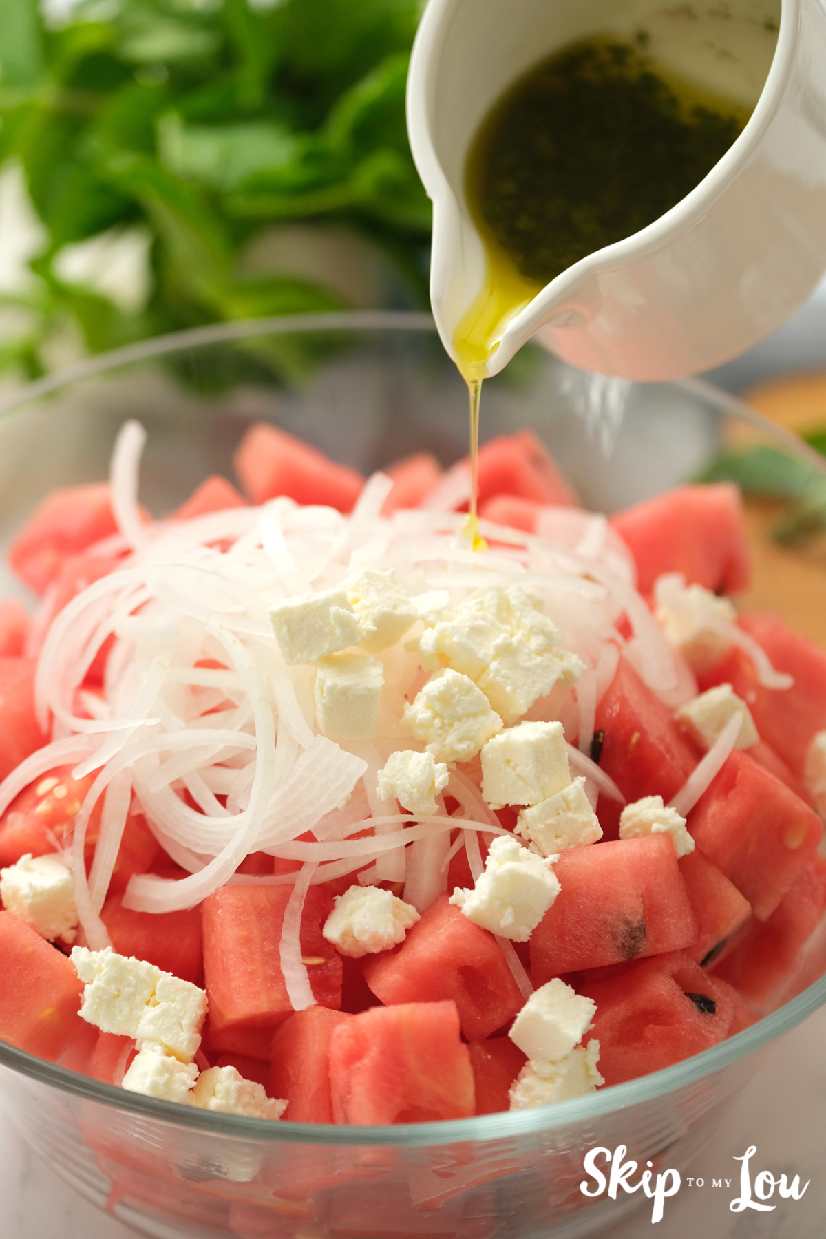 Watermelon cubes, feta cheese crumbles, onion rings and dressing ready to be tossed together, by Skip to my Lou.