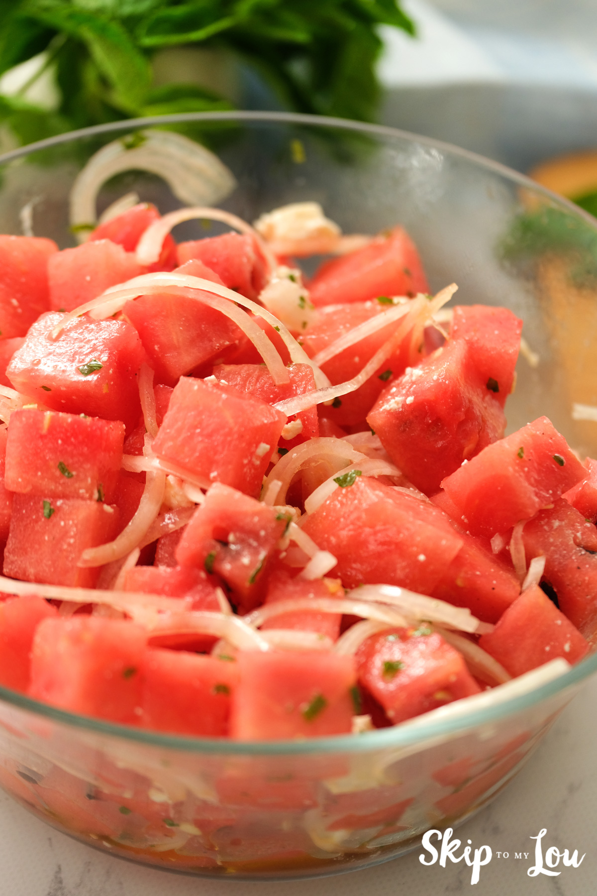Red cubes of watermelon tossed with onions, feta cheese and dressing in a clear glass bowl, by Skip to my Lou.