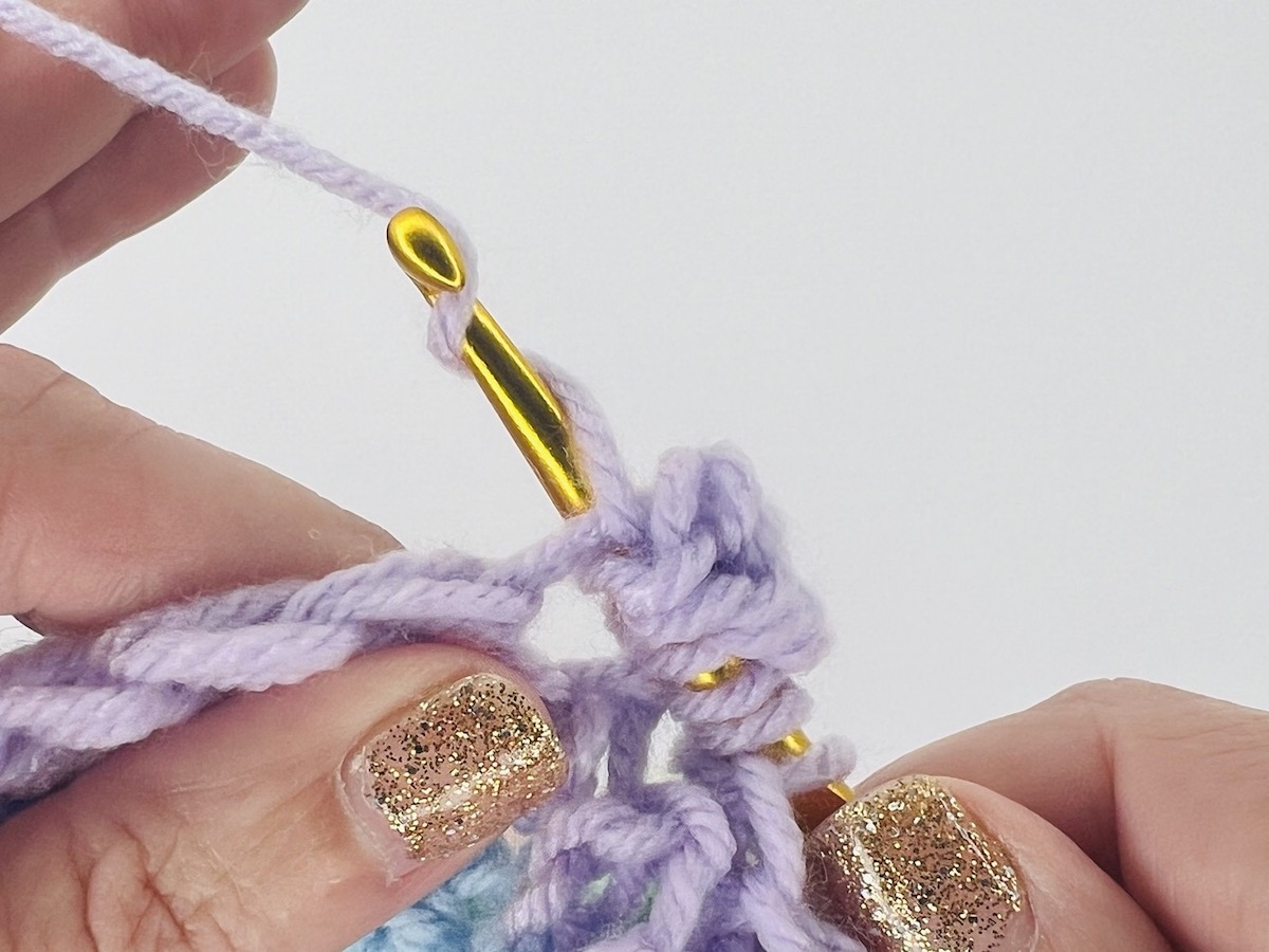 Crocheting a bobble stitch with a lot of loops on a gold hook of purple yarn.