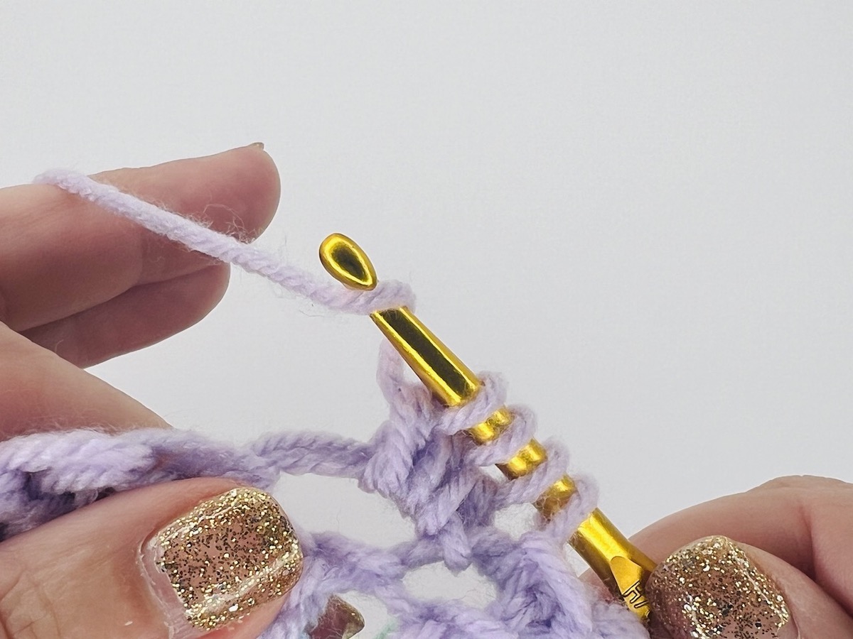 Crocheting a bobble stitch with four loops on a gold hook of purple yarn.