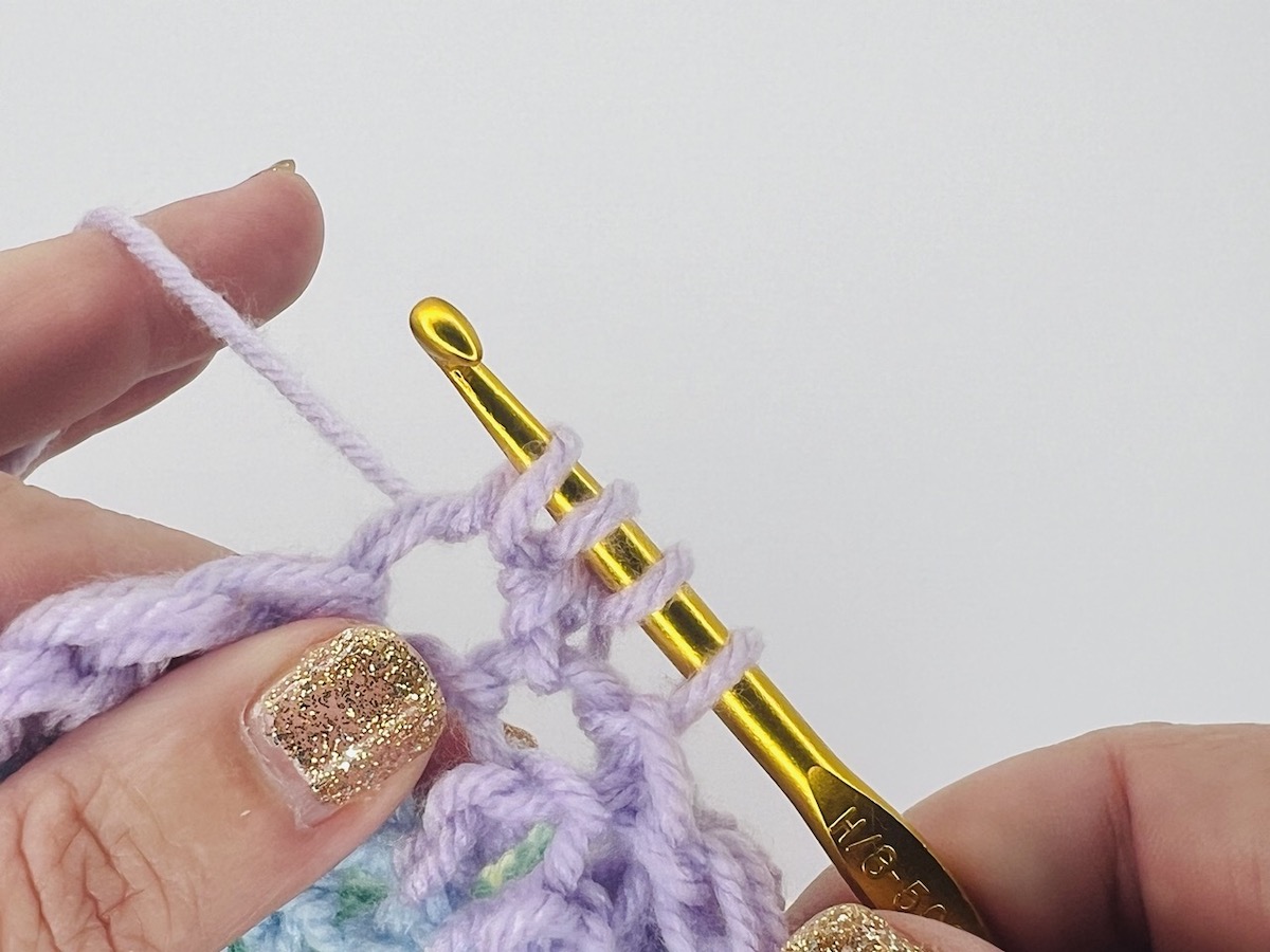Crocheting a bobble stitch with four loops on a gold hook of purple yarn. 