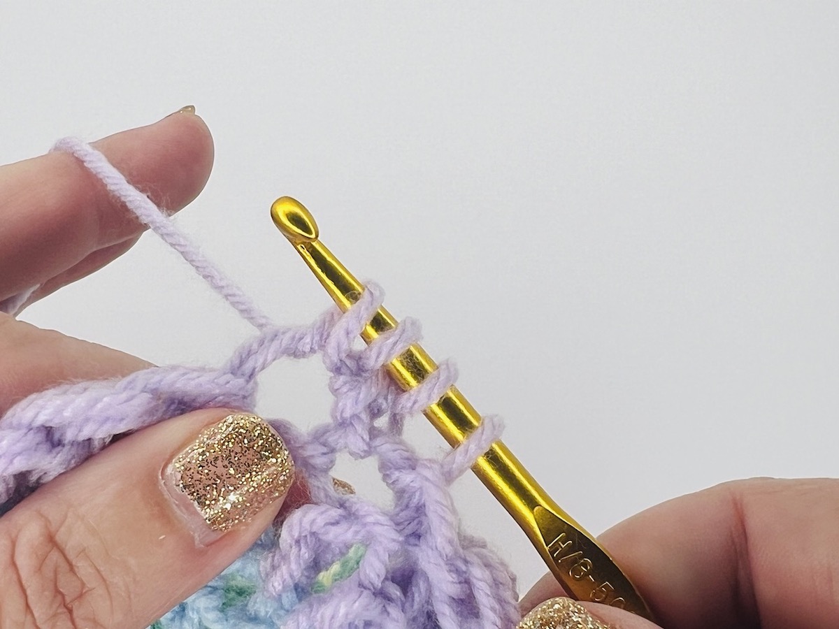 Crocheting a bobble stitch with four loops on a gold hook of purple yarn.