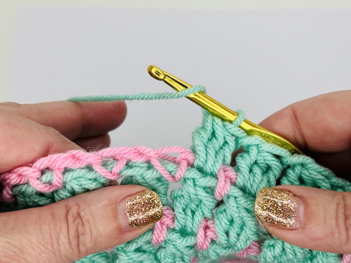 Mint green and light pink crochet cluster stitches with a gold crochet hook on a white background.