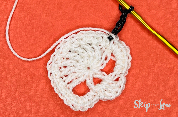 White yarn crocheted into a a skull with a black border starting on the skull on an orange background with the beginning of a black border.  