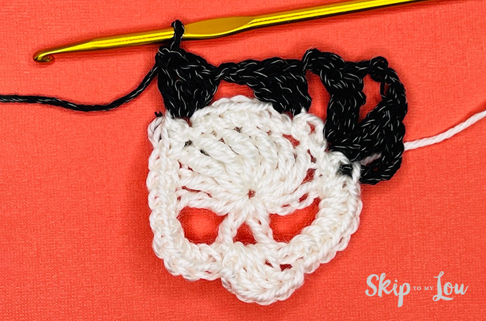 White yarn crocheted into a a skull with a black border starting on the skull on an orange background with the beginning of a black border.