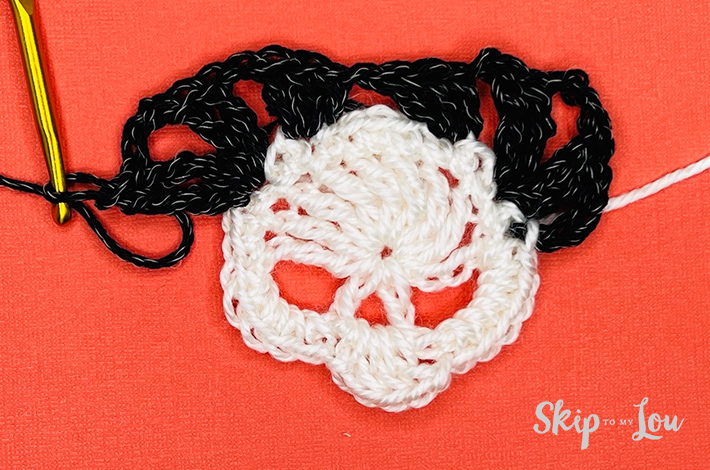 White yarn crocheted into a a skull with a black border starting on the skull on an orange background with the beginning of a black border.