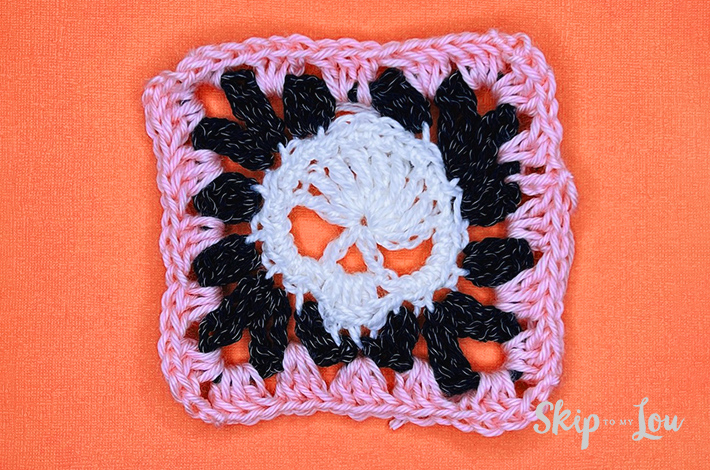 White yarn crocheted into a a skull with a black border around it and then a pink border around it.