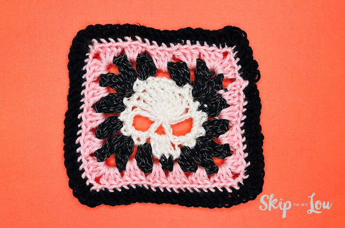A closeup of a white crocheted skull in the middle of a a pink and black granny square. 