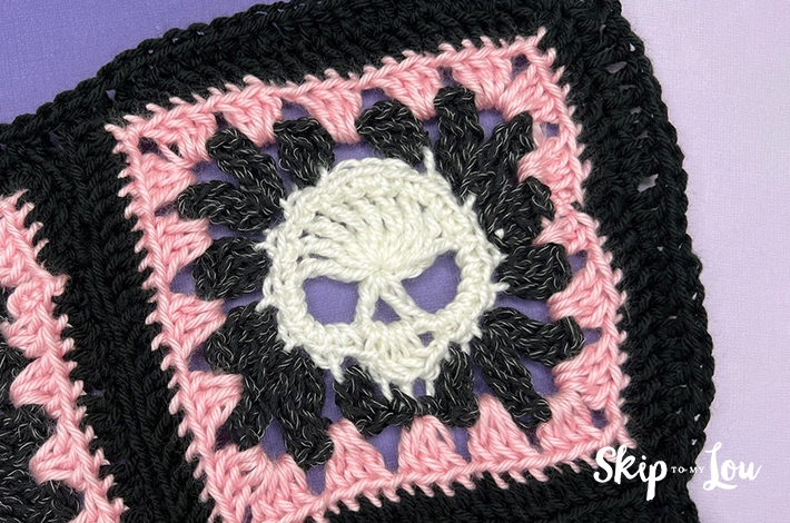 A closeup of a white crocheted skull in the middle of a a pink and black granny square.