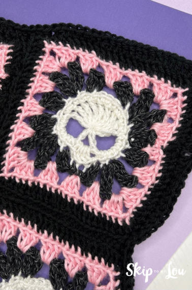 A closeup of a white crocheted skull in the middle of a a pink and black granny square.