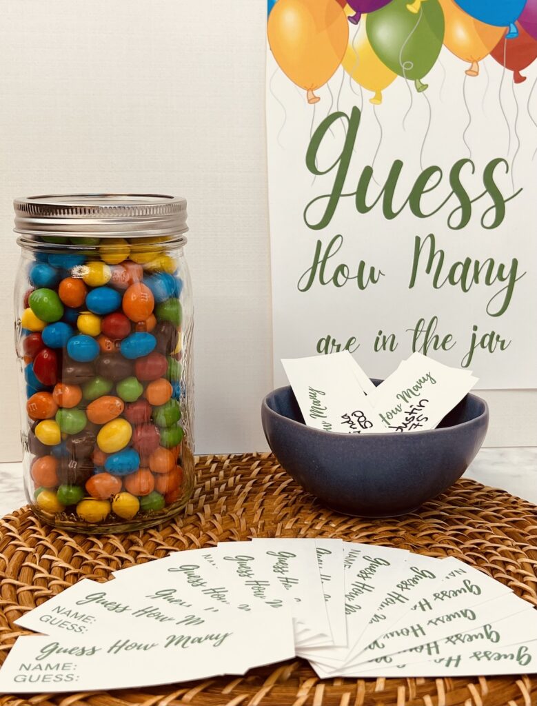 A mason jar filled with peanut M&Ms sitting on a table next to a bowl filled with cards where you can place your guess. You can download these from Skip to my Lou.