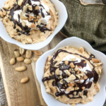 Image shows two bowls with chocolate peanut butter mousse parfait on top of a wooden board and a spoon next to it. There are loose peanuts on the table.