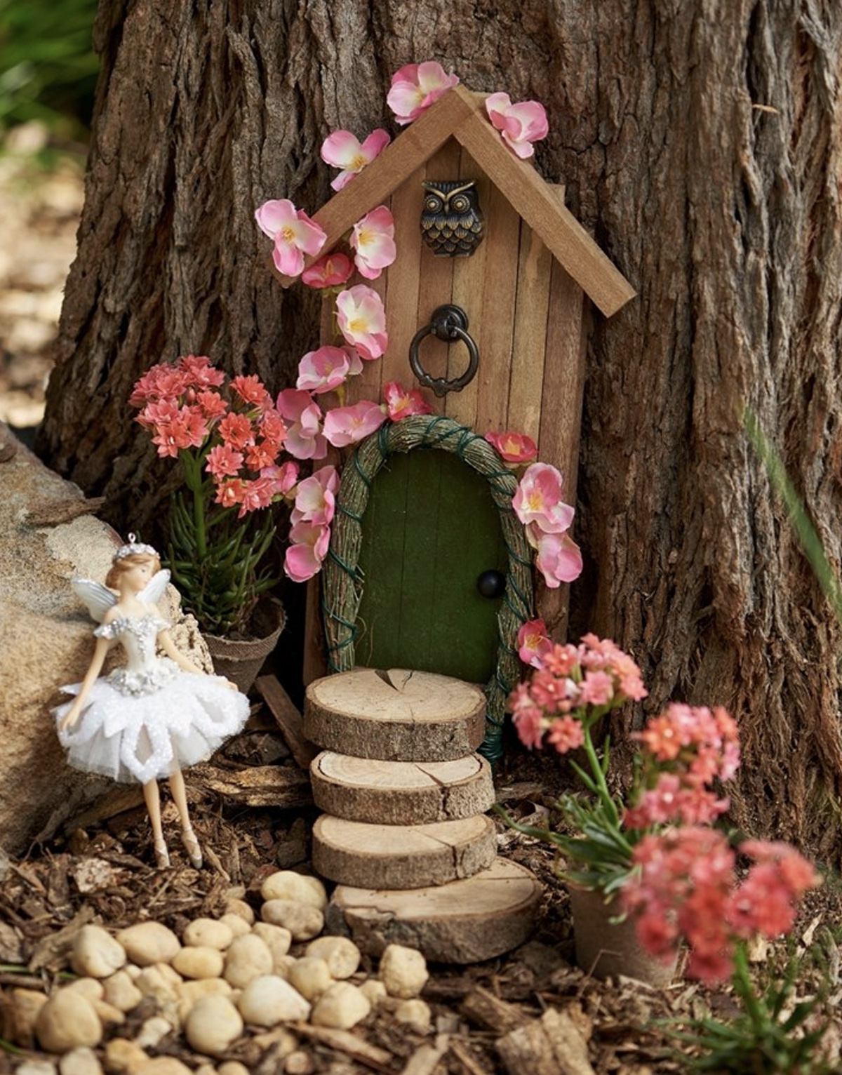 A fairy door placed against a tree trunk with circular wooden logs acting as a pathway to the door. A beautiful fairy stands outside amongst pebbles and flowers.  