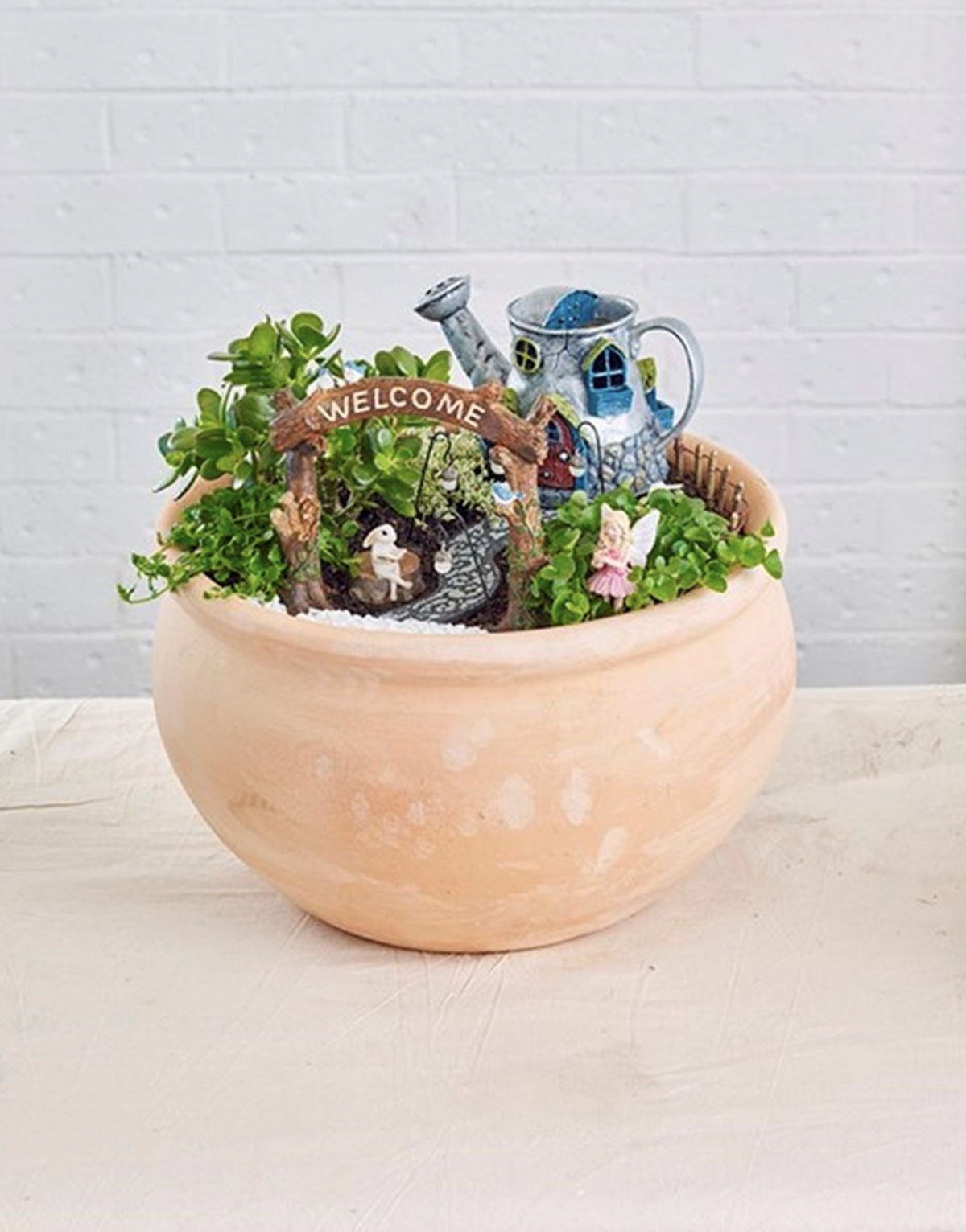 A fairy garden in a clay pot with a welcome sign, a rabbit sitting gon a stone, a fairy, and a path leading up to a fairy house, which is a tea pot. 