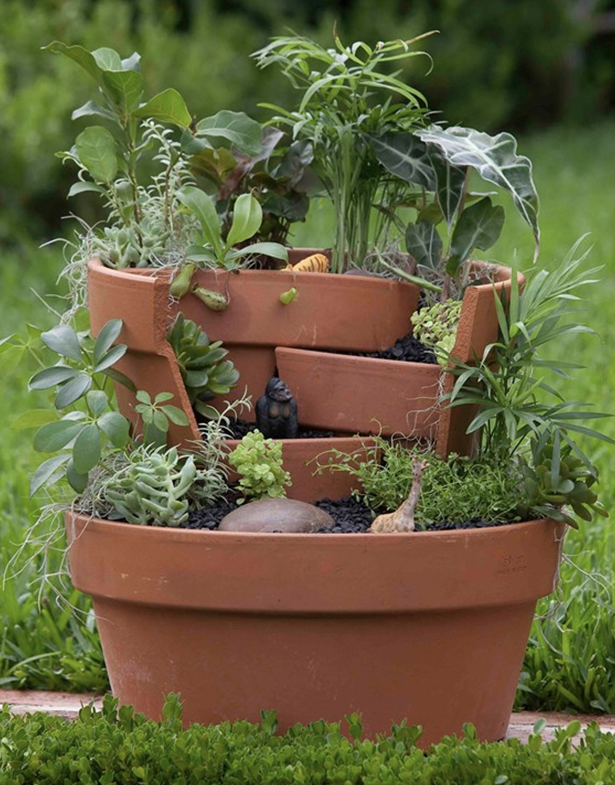 A layered fairy garden of four levels made from garden pots. The levels include various flowers, trees, and shrubbery, as well as a giraffe, gorilla, and tiger figurine that sits in the top section with taller leaves, which look like a jungle.