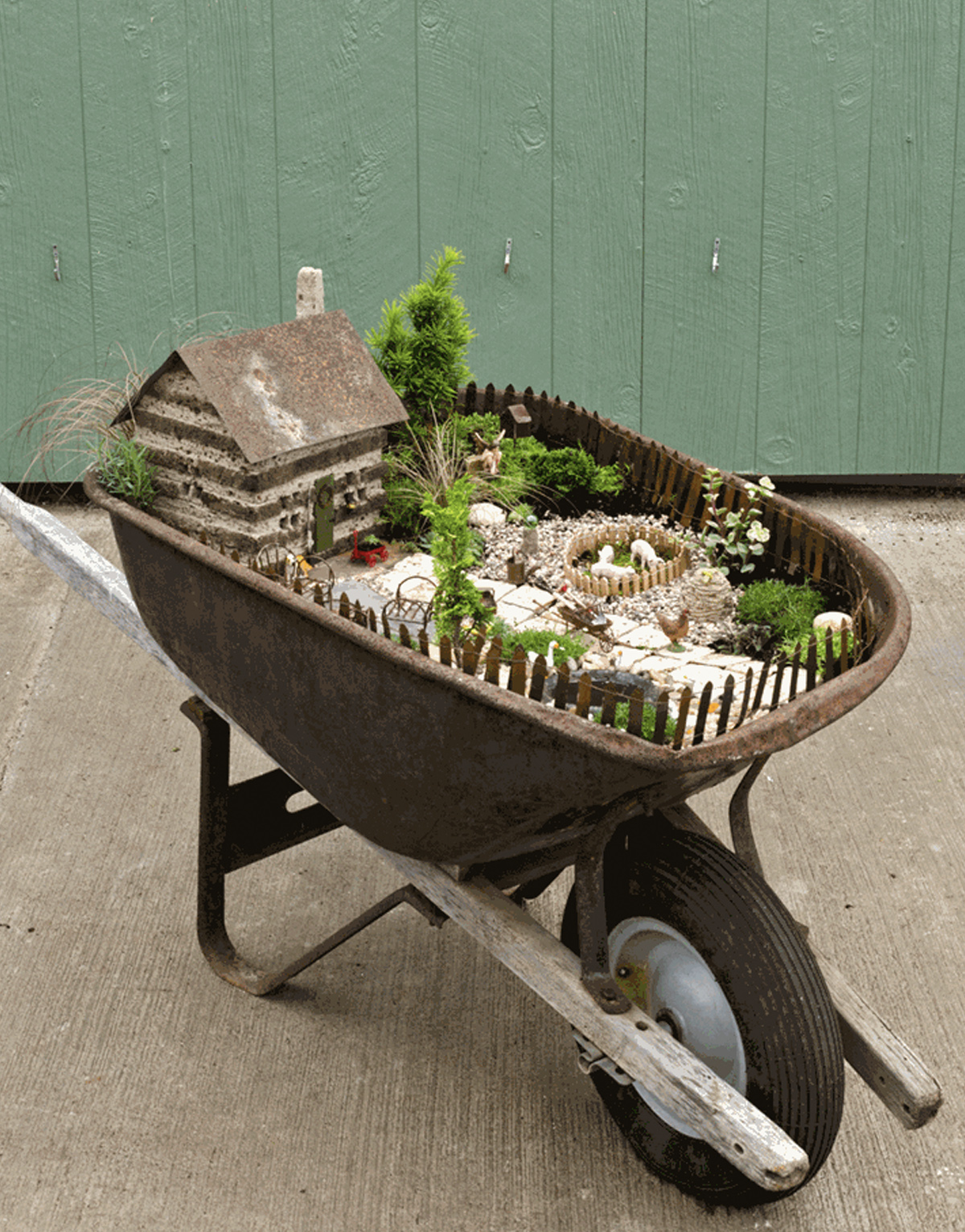 A fairy garden, featuring various flowers and other plants constructed in a wheelbarrow. It also includes a wooden fence around the perimeter and a large fairy house. 