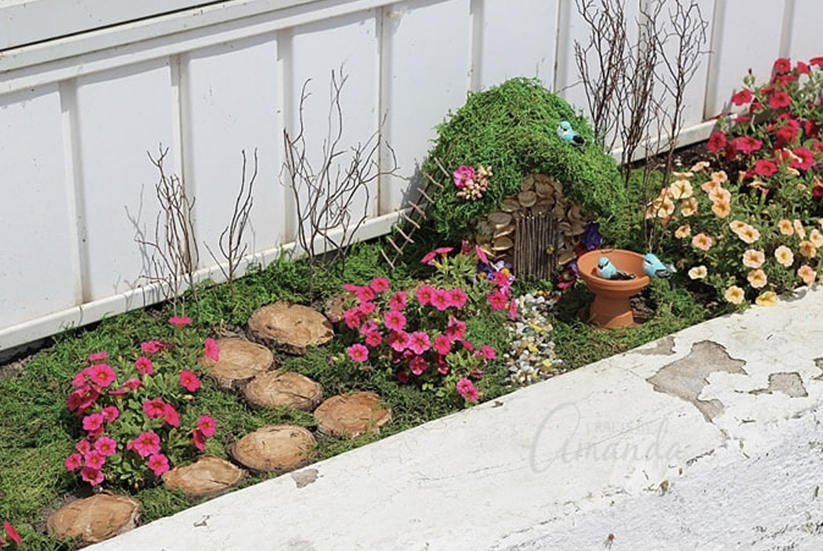 A beautiful fairy garden featuring a path made of circular log pieces, flowers, small bushes, a bird bath, and a tiny house with a moss-covered roof. 