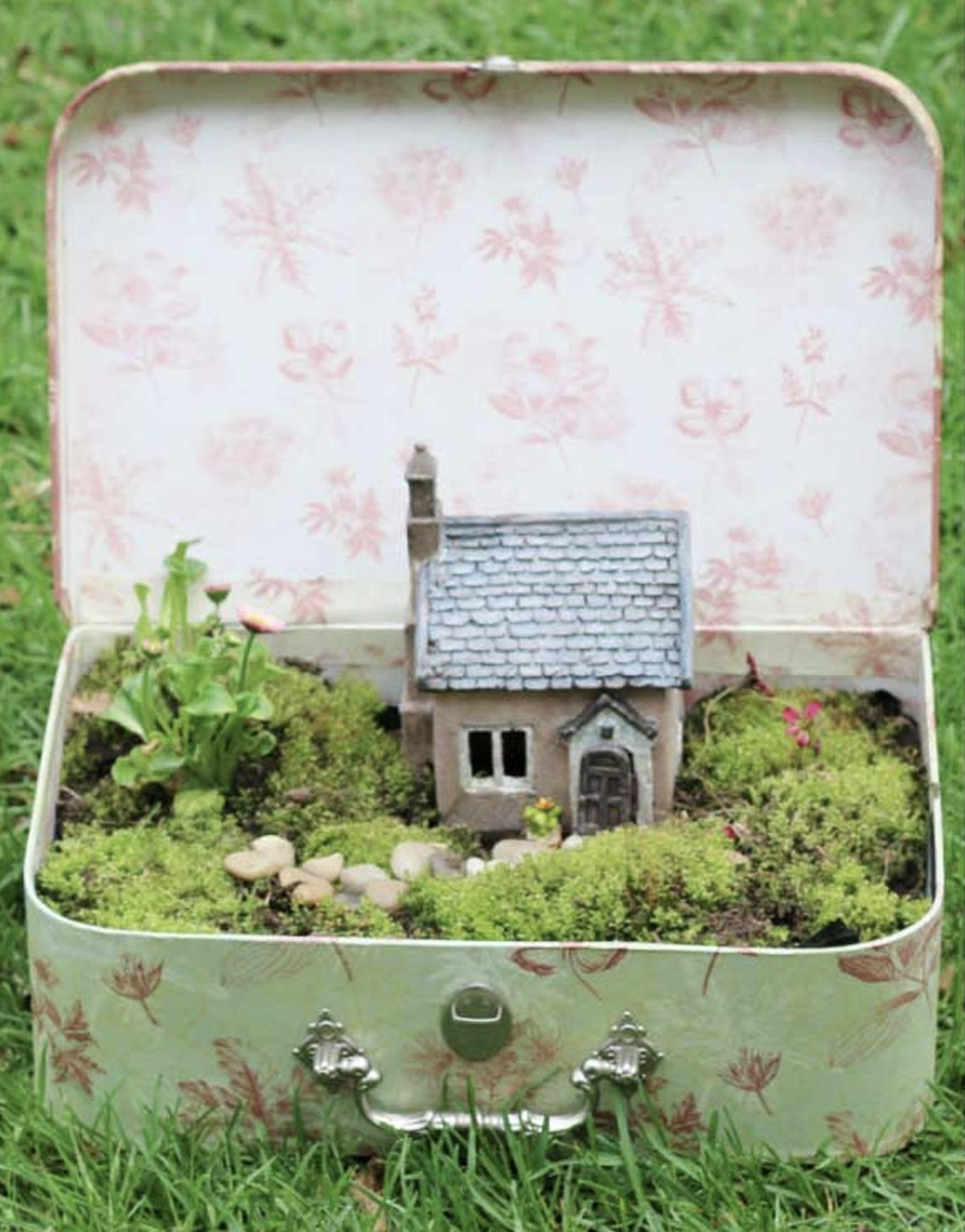 A simple fairy garden consisting of moss, bushes, a plant, cobble stones, and a simple fairy house. 