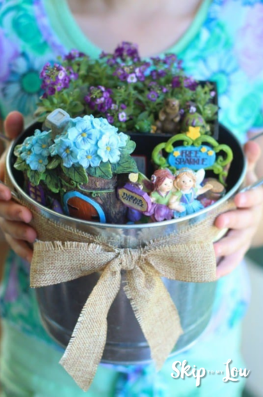 A woman is holding a bucket with a fairy garden- it has different flowers, plants, and colors. The bucket is decorated with a bow.