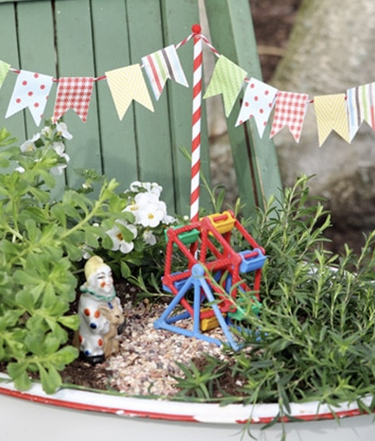 A circus fairy garden made in a garden pot which includes a Ferris wheel, a clown, two plant types, white flowers, and circus bunting. 