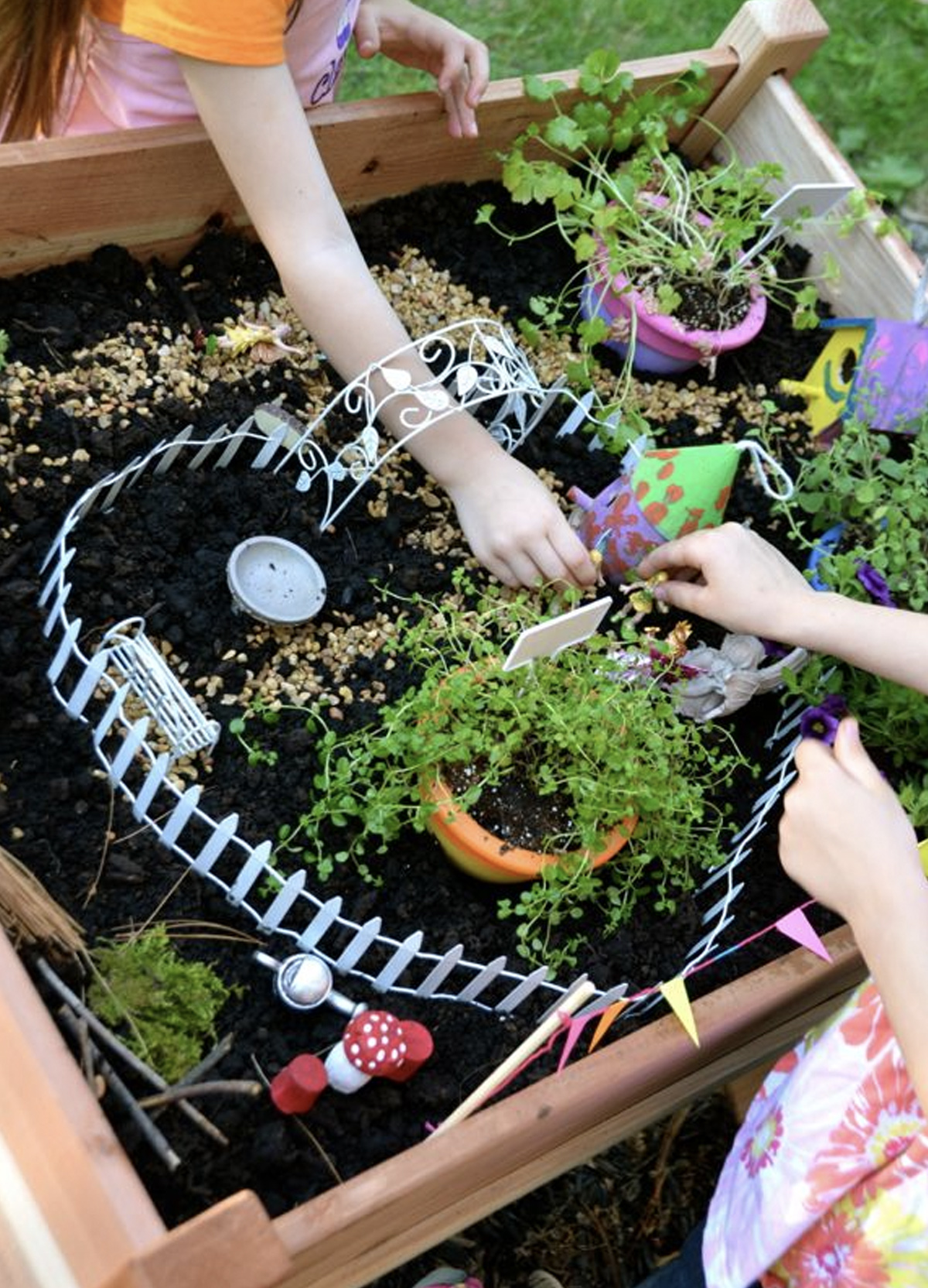 An adult and a child making a fairy garden together. It includes a soil-based floor, plants, a homemade fairy house made out of paper, figurines, and a colorful birdhouse. 