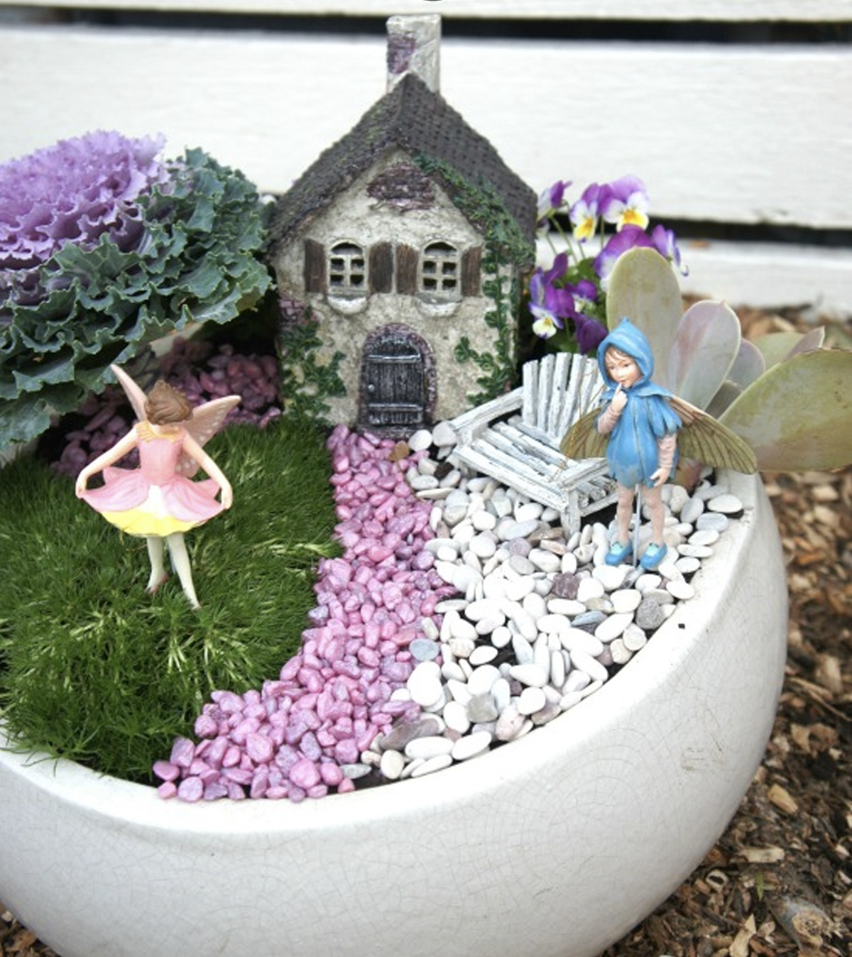 Two fairies in a simple fairy garden that includes their home, a bench constructed from wooden logs and painted white, and a pink pebble path. 
