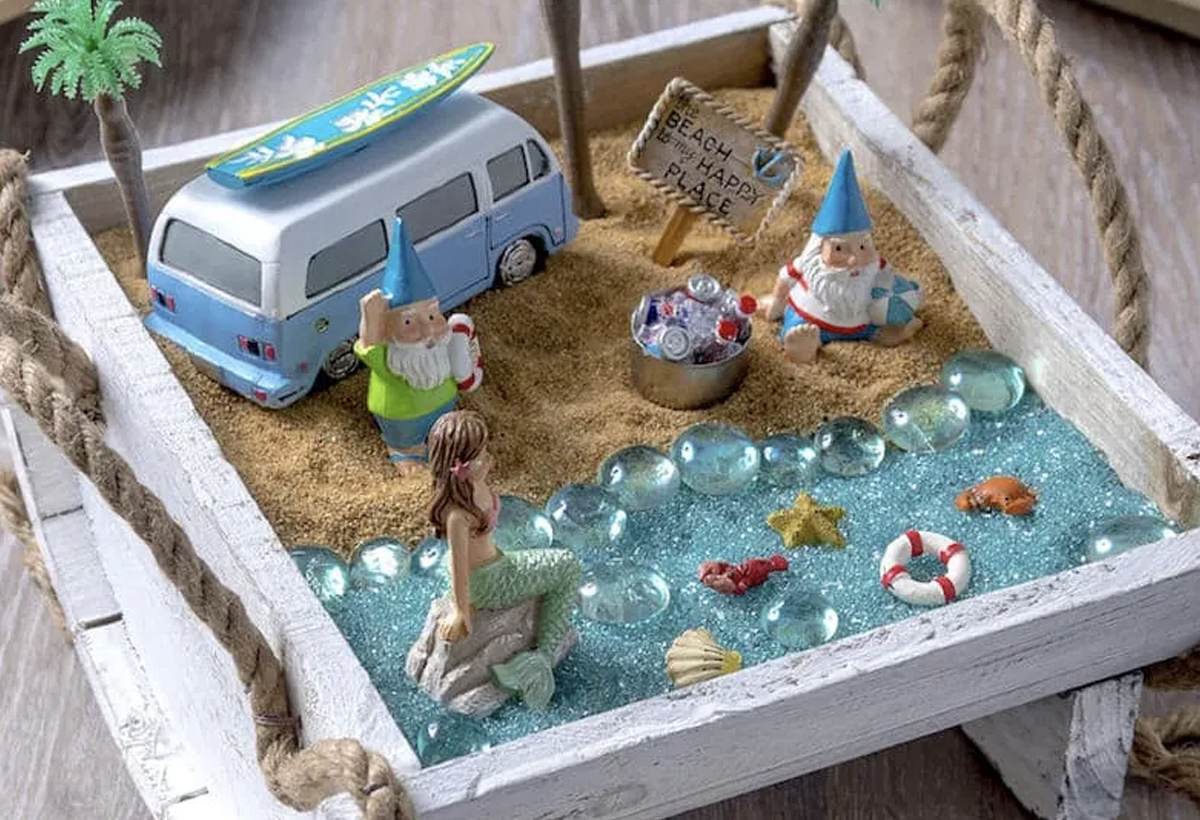 Made in a square wooden container, this fairy garden depicts a beach scene with a camper van, three gnomes, a mermaid in the water as well as sea creatures, and a life preserver. The sea itself is made from sand that has been dyed blue, with clear blue pebbles depicting waves. 