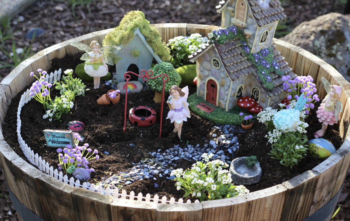 A fairy garden made in a half barrel. It features a sandy floor, flowers, a small pond, and two fairy houses, one of which has two stories and three fairies. 