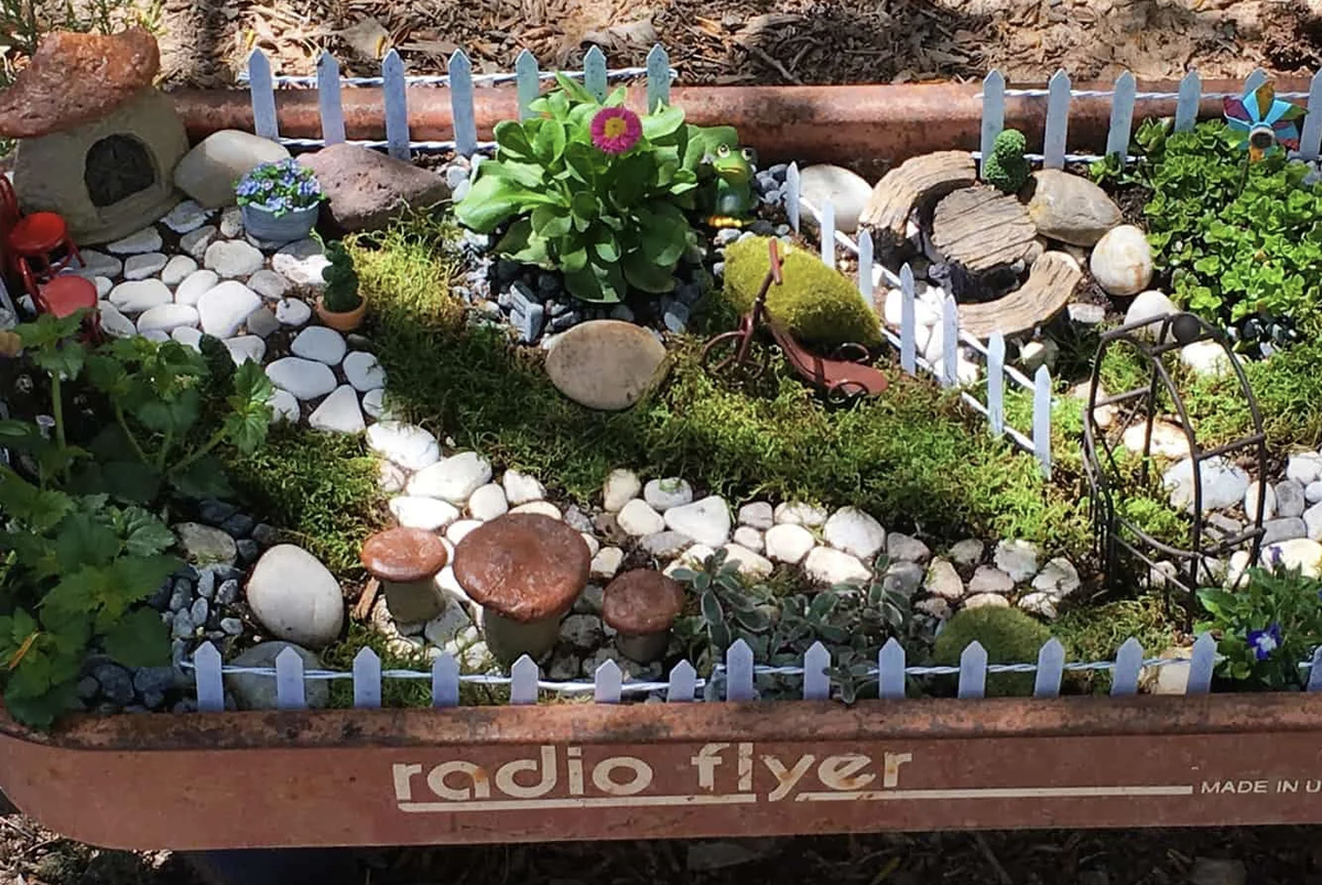 A fairy garden with a toadstool home, a white pebble path, a push bike, and a bench and table made from wood. 