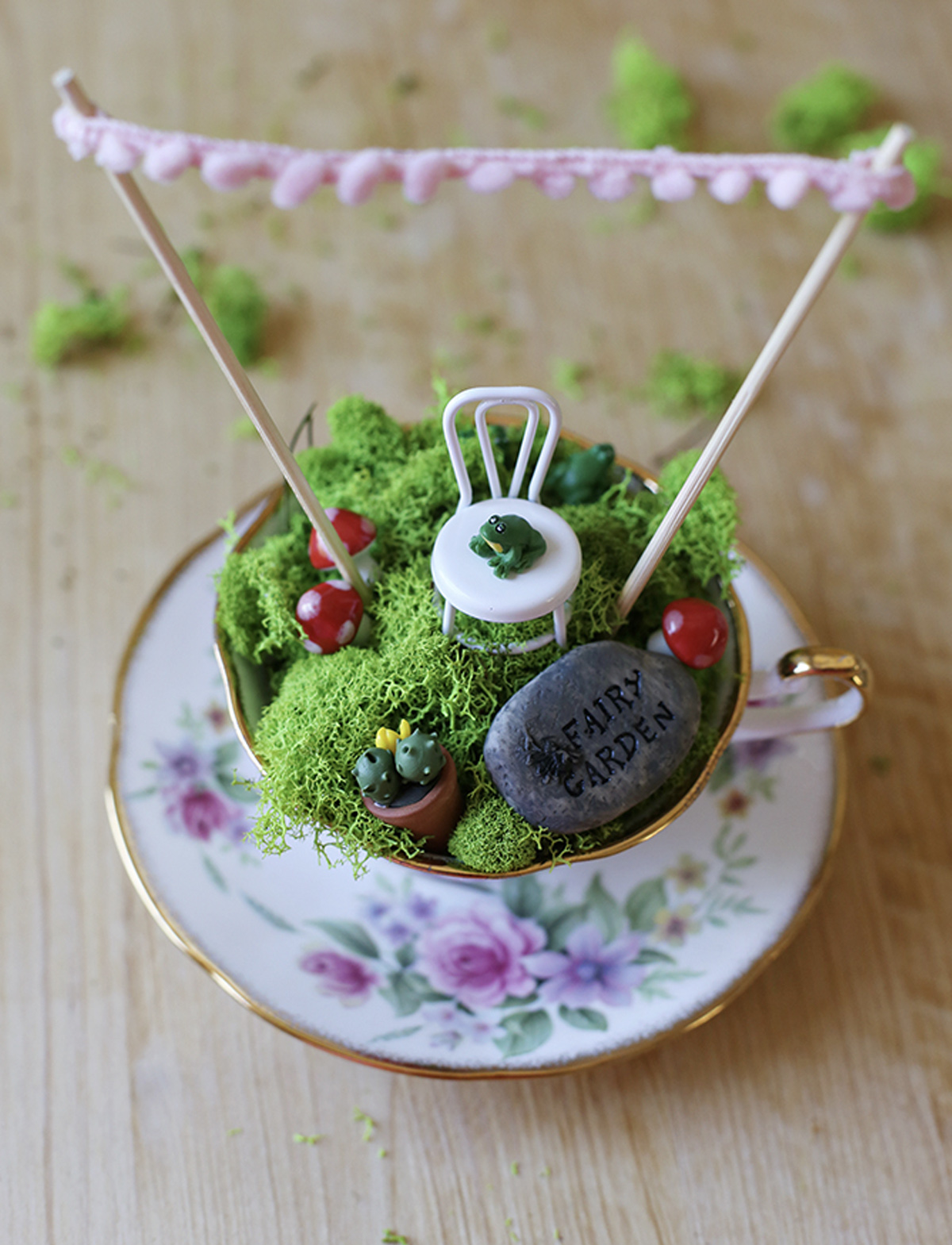 A fairy garden constructed in a teacup with moss flooring, a stone saying "Fairy Garden,” a small chair with a frog sitting on it, a pot plant, and three toadstools. 