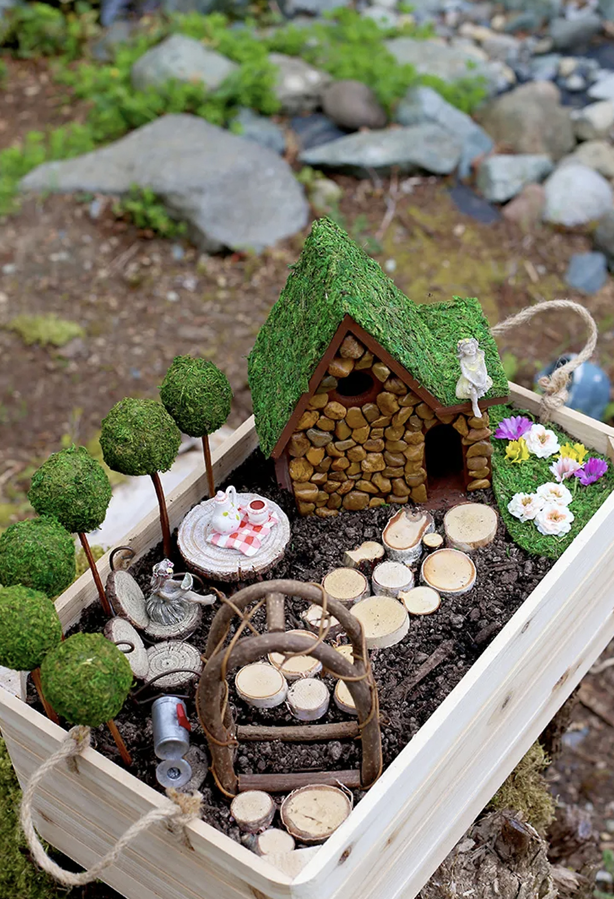 A fairy house finished with small pebbles, with a fairy sitting on its roof. The garden includes a table with a tea set, a fairy sitting on a log chair, a letter box, flowers, and a wooden archway. 