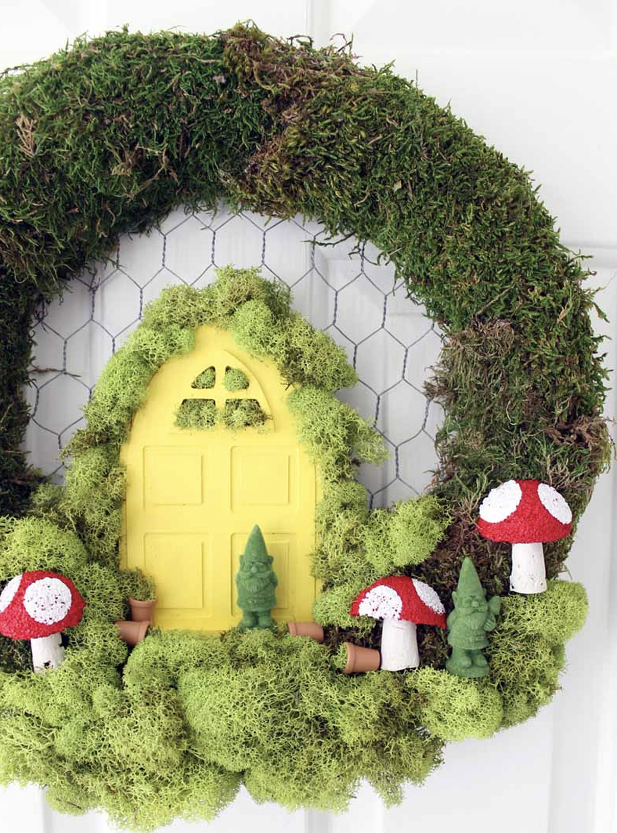 A fairy garden made within a wreath that includes a simple door, gnomes, small pot plants, toadstools, and moss. 