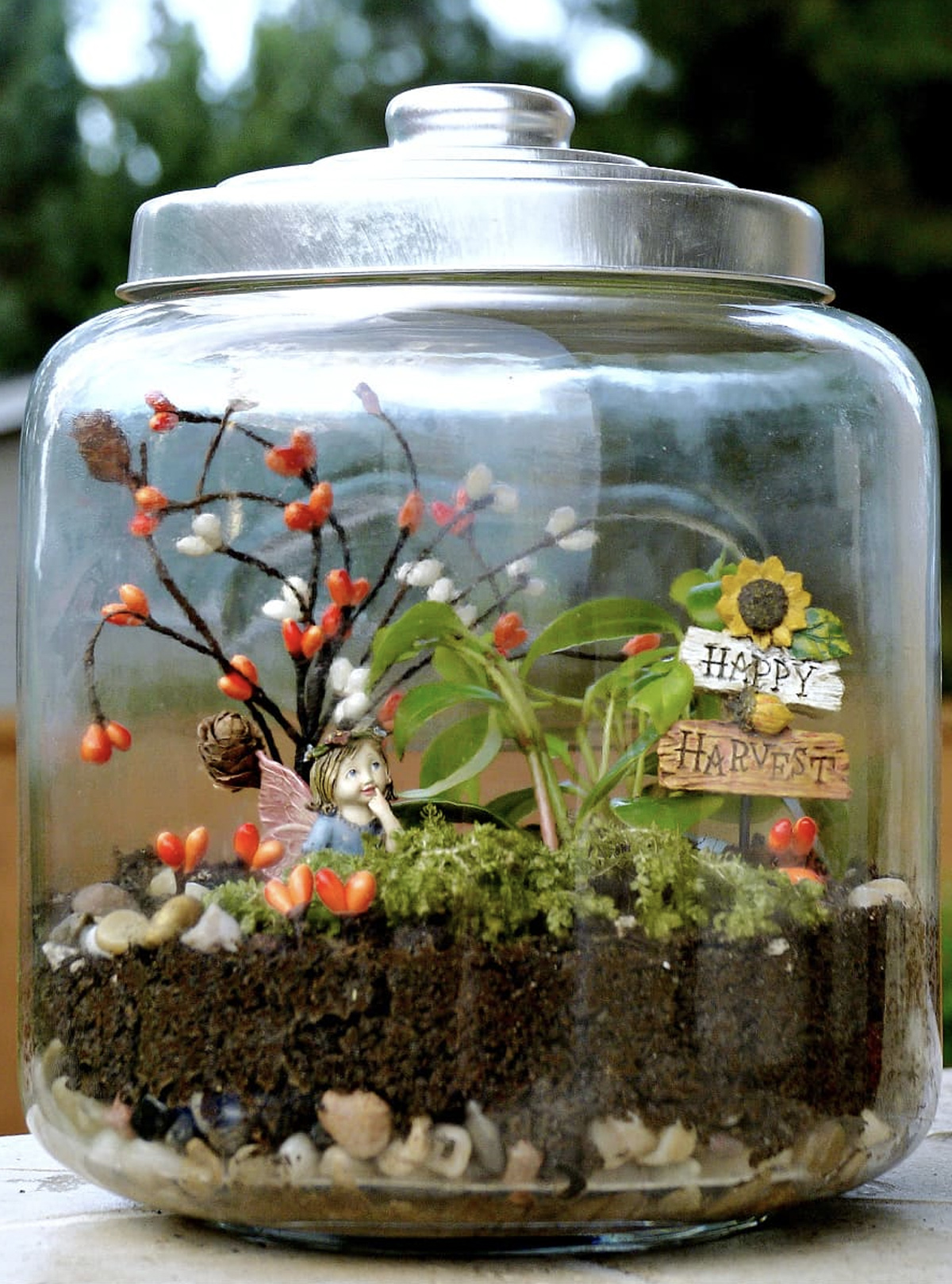 A fairy garden made within a terrarium. It includes a Happy Harvest sign, moss, soil, a tree with heart-shaped fruit, and a fairy sitting in the garden. 