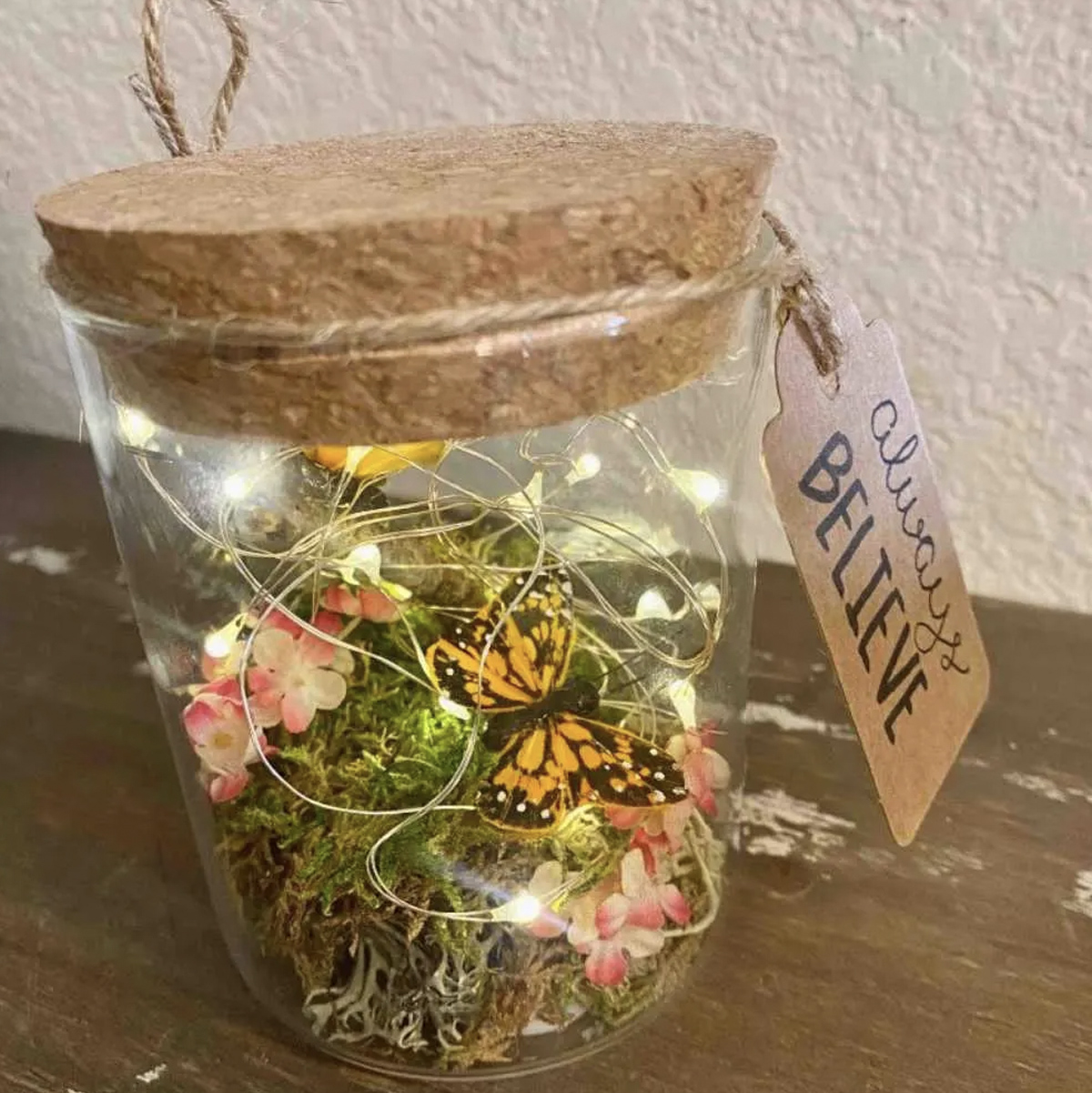 A fairy garden terrarium made in a small glass jar with moss, flowers, a butterfly, and fairy lights. 