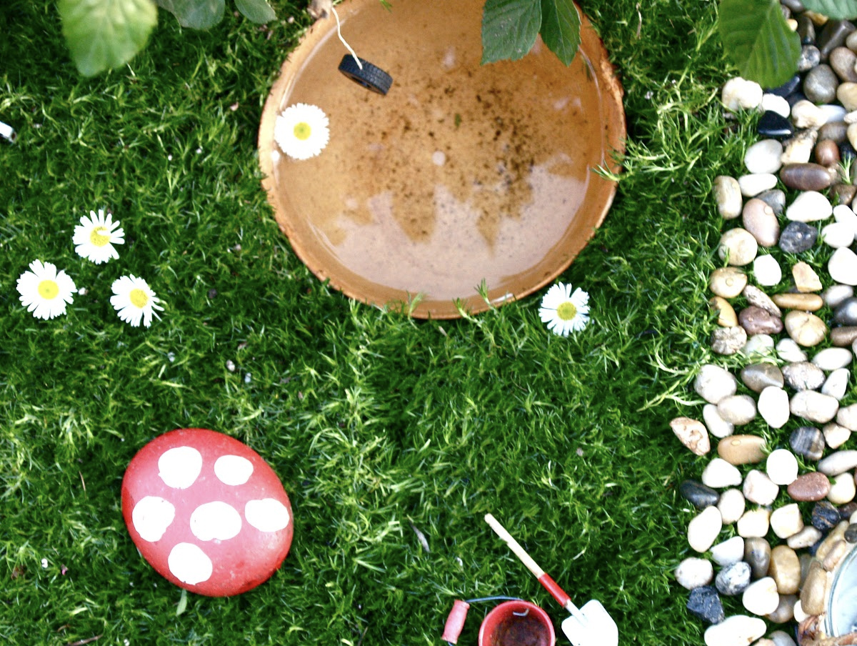 A simple fairy garden for kids with pebbles, grass, a painted toadstool made from a rock, a small bucket and spade, a clay pond, and daisies. 