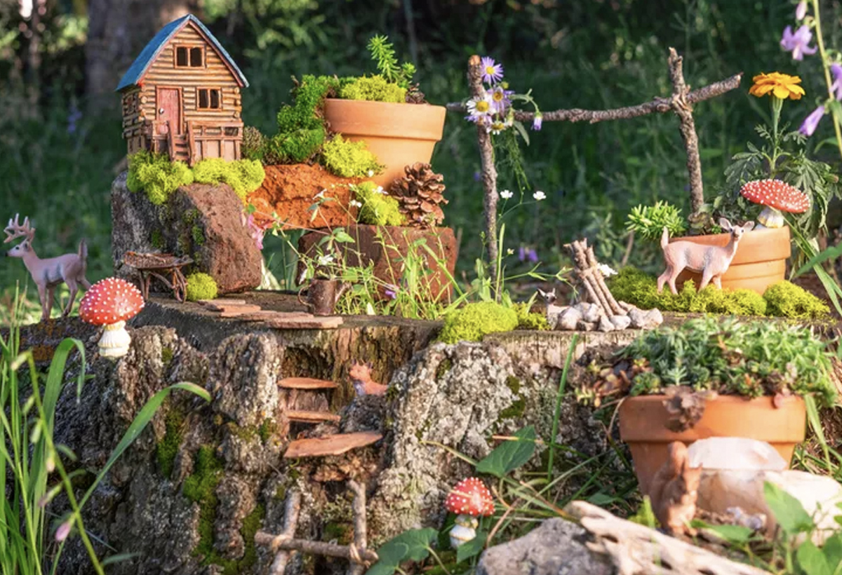 A complex layered fairy garden includes a timber log fairy house at the very top. There are toadstools, various plants, two deer, and a small wheelbarrow. 