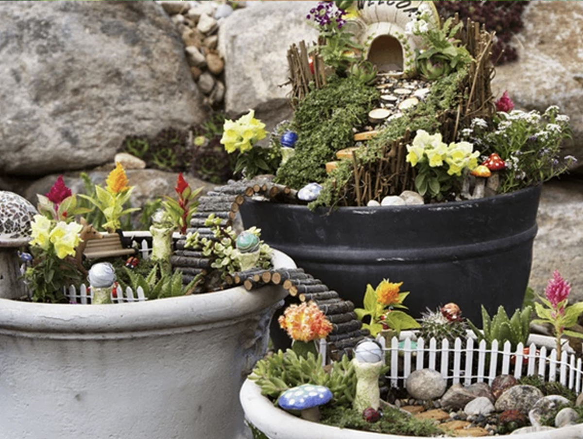 A fairy garden consisting of three separate garden pots, each with its own unique design. They are all connected by a log bridge, with the top garden including a fairy house, the middle garden a bench, and the bottom garden, a toadstool. 