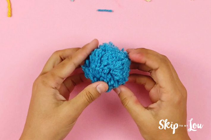 The final product. A blue pom pom held by two hands over a pink background.