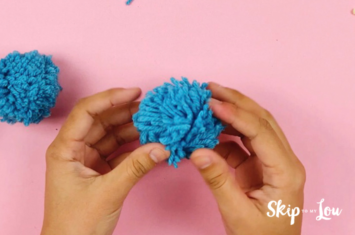 The perfect pom pom. A blue pom pom held by two hands over a pink background.