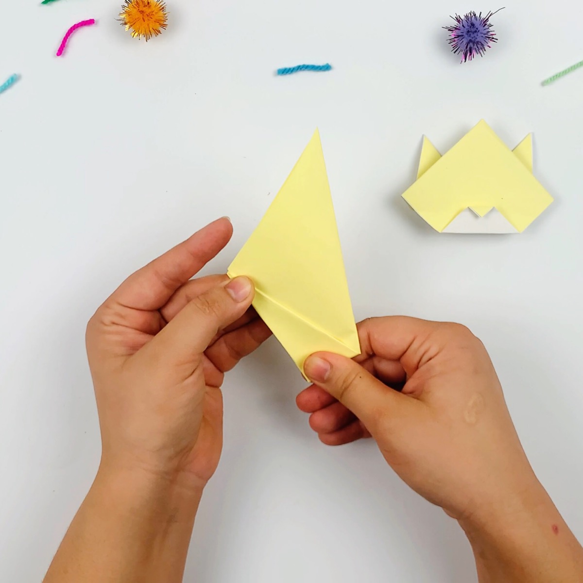 The first section of the origami cat's tail is folded by taking the bottom corner and folding it along the straight line of the paper towards the top left.