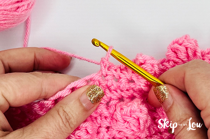 Two hands holding a crochet hook and working with pink yarn on the first step of the popcorn stitch.