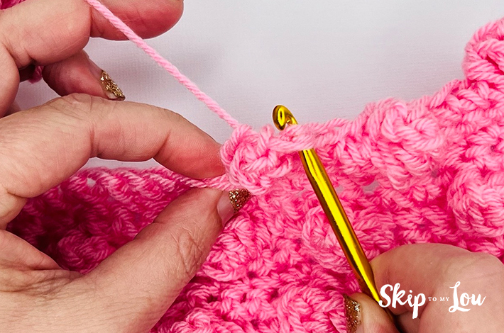 Two hands holding a crochet hook and working with pink yarn on the 10th step of the popcorn stitch.