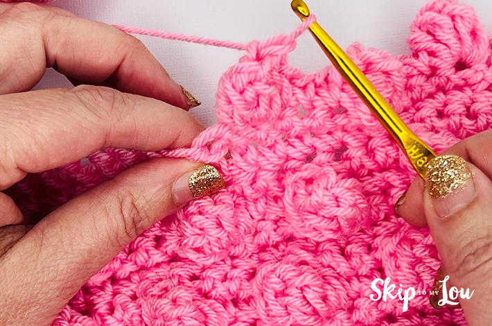Two hands holding a crochet hook and working with pink yarn on the 11th step of the popcorn stitch.
