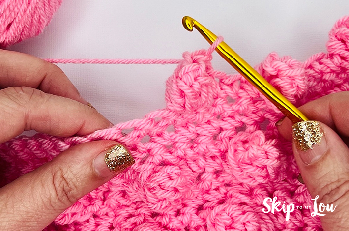 Two hands holding a crochet hook and working with pink yarn on the 12th step of the popcorn stitch.