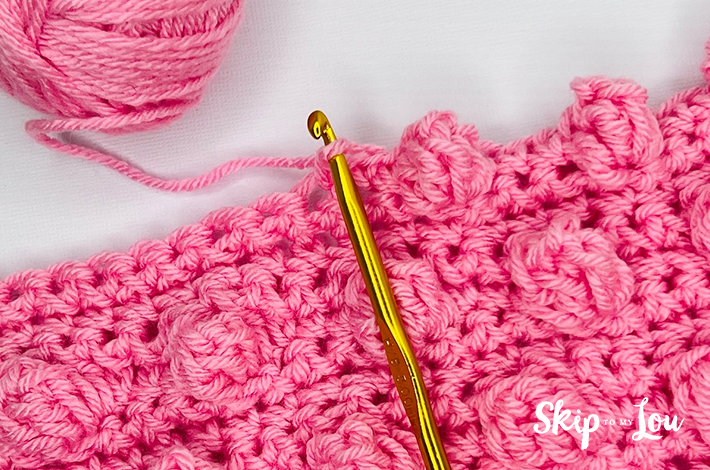 Two hands holding a crochet hook and working with pink yarn on the 13th step of the popcorn stitch.