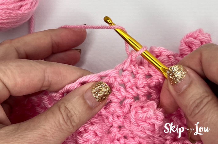 Two hands holding a crochet hook and working with pink yarn on the second step of the popcorn stitch.