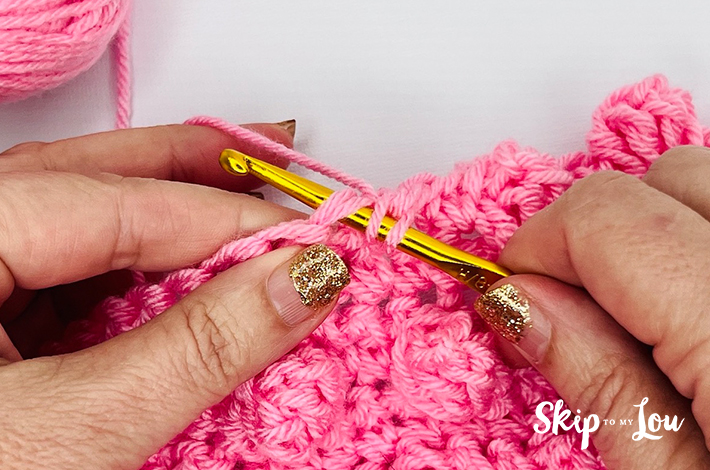 Two hands holding a crochet hook and working with pink yarn on the third step of the popcorn stitch.