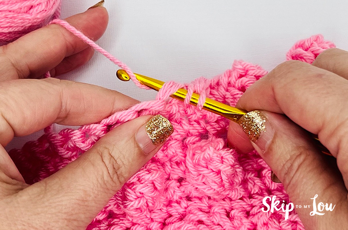 Two hands holding a crochet hook and working with pink yarn on the fourth step of the popcorn stitch.