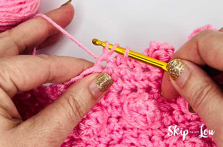 Two hands holding a crochet hook and working with pink yarn on the fifth step of the popcorn stitch.
